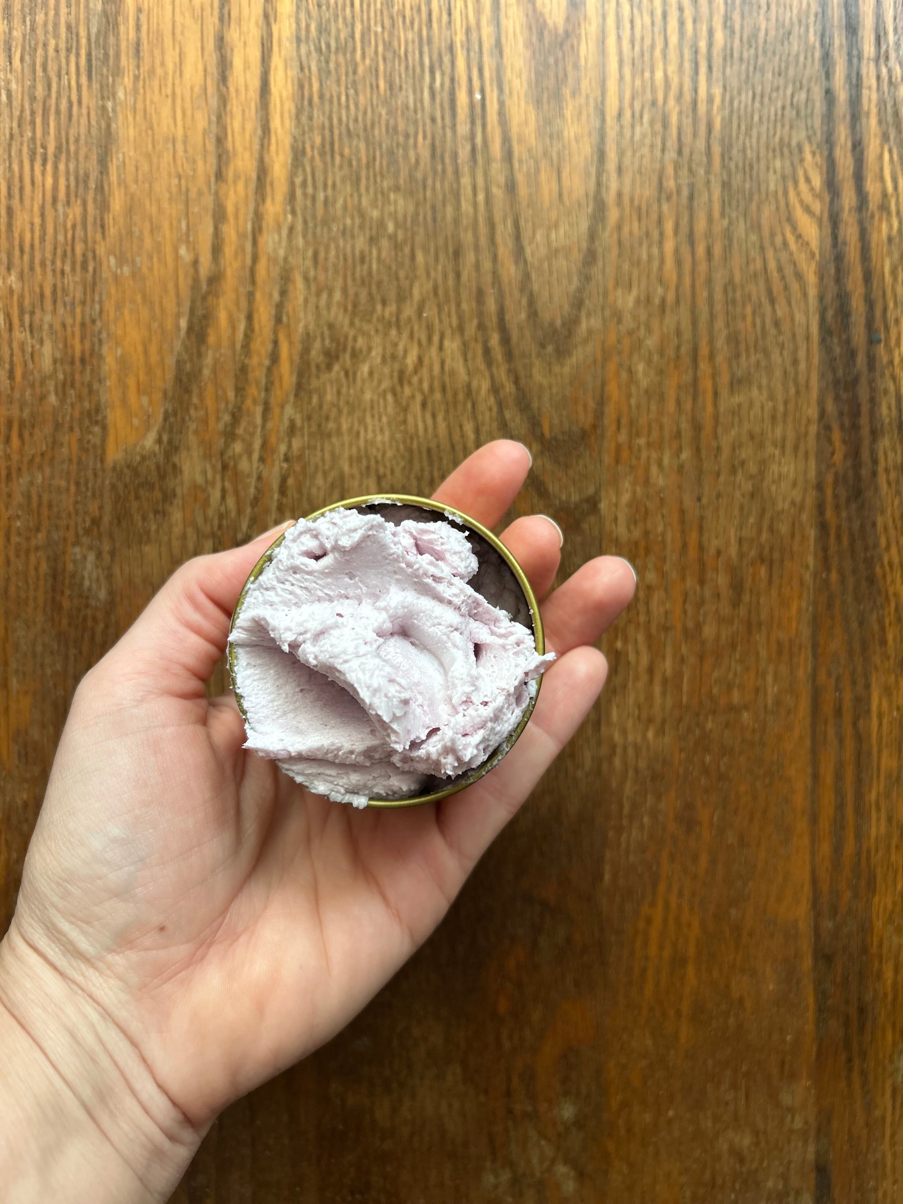 A hand holding an open tin of soft pink whipped tallow balm